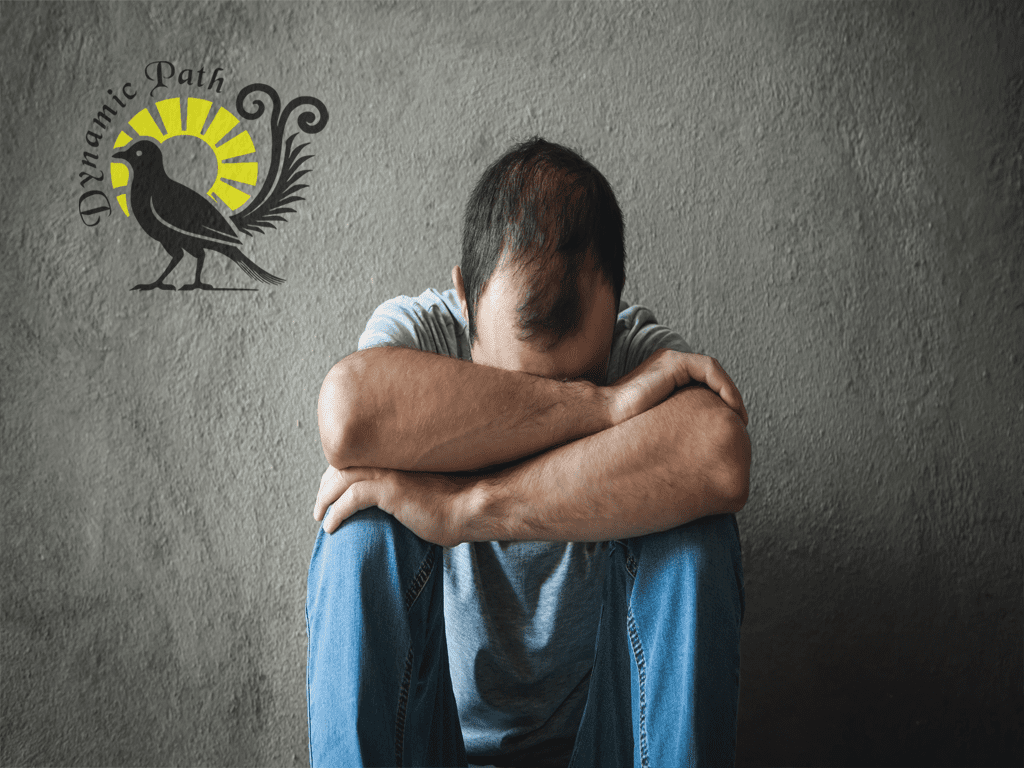 Person sitting with head down against gray wall beside Dynamic Pathways logo with lyre bird and sunburst